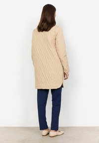 Beige vadderad lång jacka med ett texturerat mönster, matchat med marinblå byxor och ljusbeige platta skor. Bakåtsikt av outfiten.