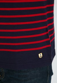 Camisa a rayas azul marino con rayas horizontales rojas y un pequeño emblema de ancla amarilla bordado en la esquina inferior izquierda.