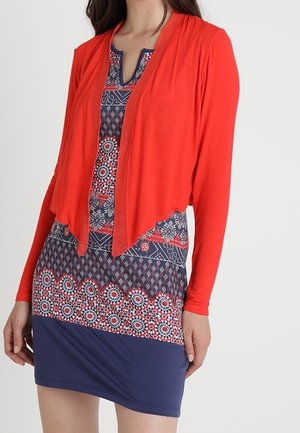 Mujer vestida con un vestido azul y rojo con un diseño floral y geométrico, combinado con un cárdigan rojo brillante abierto al frente.