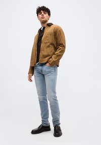 Νεαρός άνδρας στέκεται μπροστά σε λευκό φόντο, φοράει καφέ μπουφάν, μαύρο πουκάμισο, ανοιχτό μπλε τζιν και μαύρα παπούτσια.