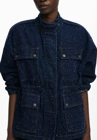Giacca di jeans blu scuro con superficie testurizzata, colletto alto, due tasche sul petto e bottoni in tonalità argento. Vestibilità leggermente oversize.