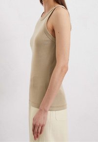 Mujer vestida con una blusa sin mangas beige ajustada y pantalones color crema, vista de perfil sobre un fondo blanco liso.