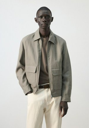Uomo con giacca beige, camicia marrone, pantaloni crema e cintura marrone, in piedi davanti a uno sfondo chiaro uniforme.