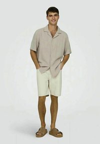 Lys beige kortærmet skjorte med knapper, cremefarvede shorts og brune slides. Glat stof og afslappet pasform med krave.