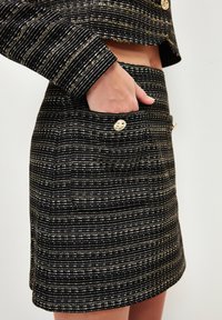Jupe en tweed à rayures noires et dorées avec une coupe ajustée, poche visible et accents en métal doré.
