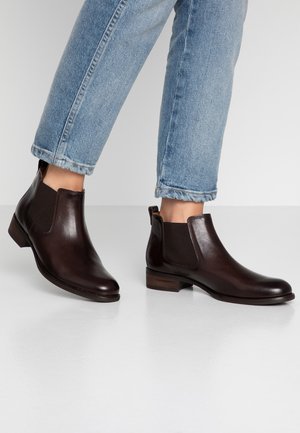 Personne portant des bottines Chelsea en cuir brun foncé, associées à un jean bleu clair retroussé, debout sur un sol blanc.