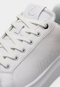 Κοντινή λήψη λευκού αθλητικού παπουτσιού με υφασμένα κορδόνια, διάτρητη επωνυμία στην πλευρά και ανάγλυφο λογότυπο στη γλώσσα.