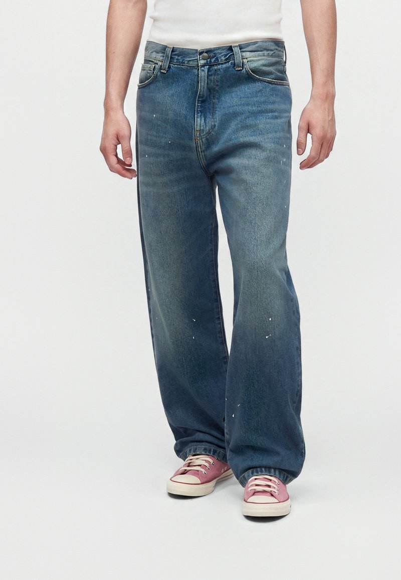 Personne portant un jean bleu délavé ample avec de petites éclaboussures de peinture blanche et des baskets en toile rose avec des lacets blancs.