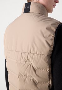 Beige polstret vest med høy krage, med horisontal søm, en svart logo-patch ved halsen, og mørkt stoff på sidene.