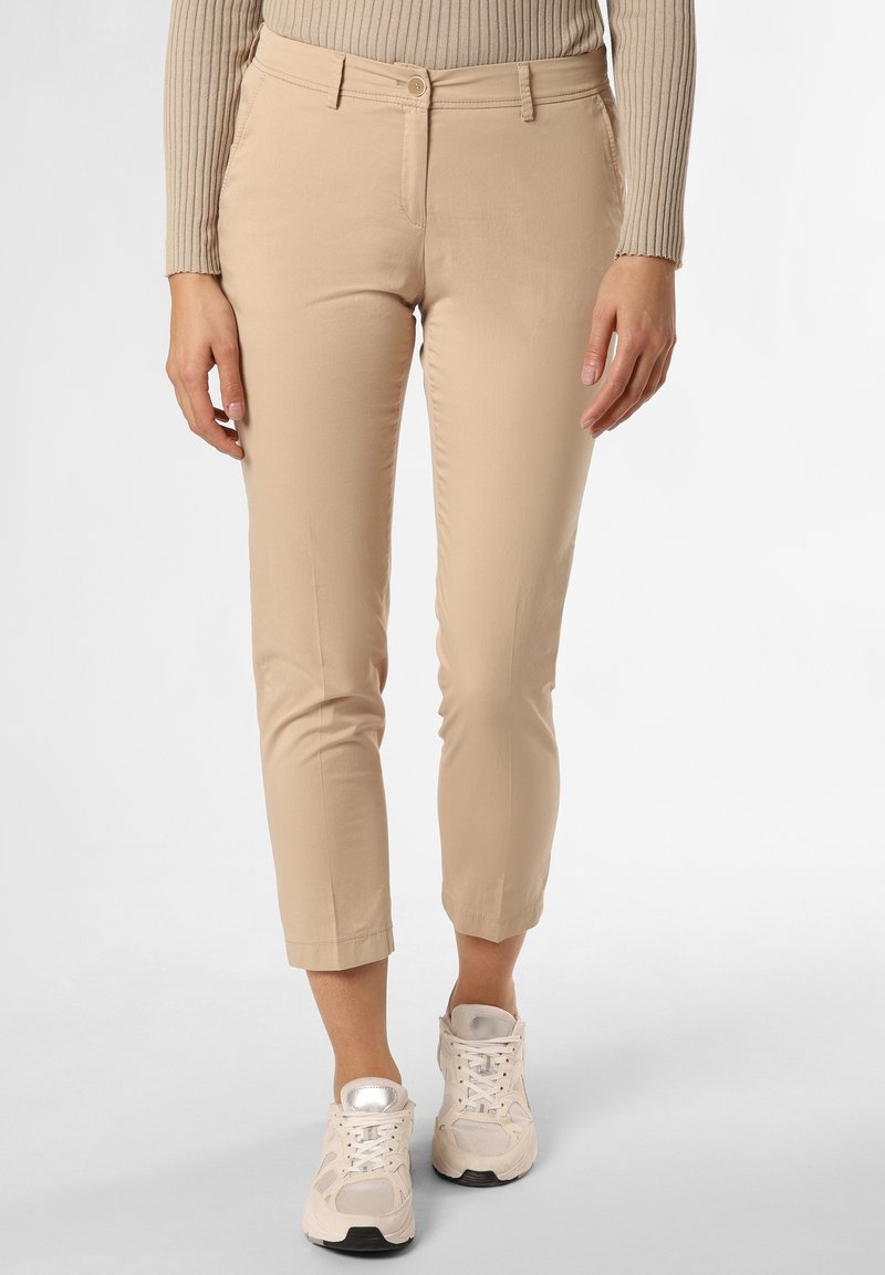 Kobieta w beżowych, krótkich spodniach, beżowym żakardowym swetrze i białych sportowych butach, stojąca na białym tle.