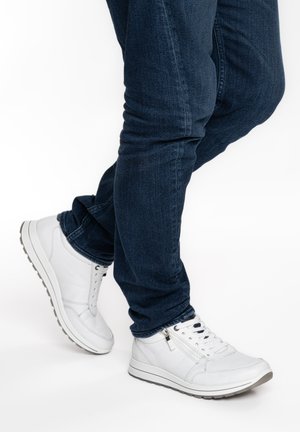 Person wearing dark blue jeans and white lace-up sneakers with side zipper, standing with one foot slightly raised.
