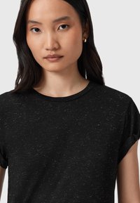 Mujer joven con cabello largo y oscuro, lleva pequeños pendientes de aro de oro y una camiseta de manga corta con manchas negras contra un fondo liso.