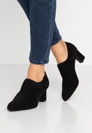 Personne portant un jean bleu foncé et des bottines en daim noir à talon moyen, debout sur un sol blanc brillant.