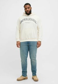 Kremowa bluza z kapturem z grafiką "JACK&JONES", w połączeniu z jasnoniebieskimi dżinsami i brązowymi butami, z kieszenią na przodzie.