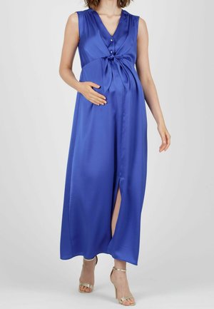 Donna incinta che indossa un vestito in raso blu reale senza maniche con scollo a V, spacco frontale e sandali dorati con tacco alto.