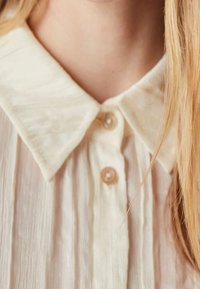 Chemisier couleur crème en tissu texturé, doté d'un col classique et de deux boutons sur le devant. Les bords du col sont délicatement arrondis.