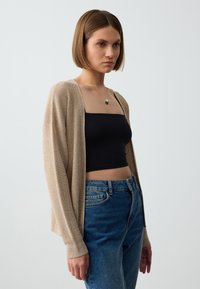Beige Strickjacke mit langen Ärmeln, lockerer Passform und gerippten Bündchen, über einem schwarzen enganliegenden Oberteil getragen und kombiniert mit blauen hoch taillierten Jeans.