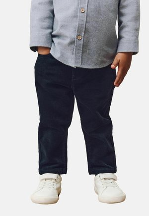 Enfant portant une chemise grise boutonnée, un pantalon en velours noir avec une main dans la poche, et des baskets blanches sur fond blanc.