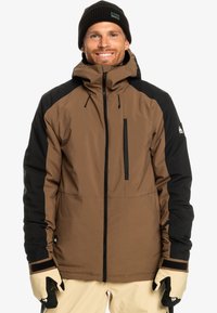 Braune und schwarze isolierte Jacke mit hohem Kragen, Reißverschlusstasche und elastischen Bündchen. Kombiniert mit beigen Schneehosen und Handschuhen.