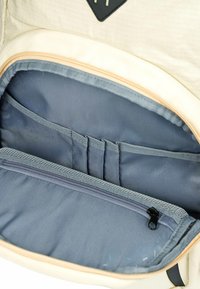 Interno di uno zaino color crema con fodera grigia, numerose tasche per riporre oggetti e un compartimento con cerniera per l'organizzazione.