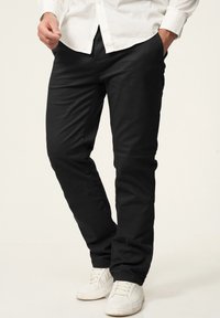 Schwarze Stretchhosen mit glatter Textur, geradem Bein Design und Seitentaschen, getragen mit weißen Sneakers und einem Hemd mit Knöpfen.