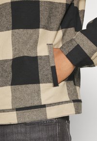 Hånd i lommen på beige og sort ternet jakke båret med mørkegrå jeans, der fremhæver stoftekstur og lommedetaljer.