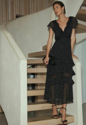 Frau in schwarzem, transparentem Polka-Dot-Kleid mit Rüschen und schwarzen Schuhen auf Holztreppe stehend, hält eine schwarze Clutch und schaut zur Seite.
