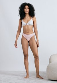 Λευκό μπικίνι τοπ με λεπτές τιράντες και αντίθετο ροζ κάτω μέρος. Λείο ύφασμα με ελάχιστες λεπτομέρειες, σε μοντέλο που στέκεται όρθιο.