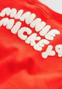 Punainen collegepaita, jossa on tekstuurista, pörröistä valkoista kirjontaa, joka sanoo "Minnie Mickey". Sujuvaa kangasta, jossa on pyöreä kaula-aukko.