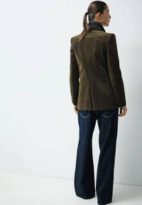 Blazer in velluto a coste verde scuro con un design strutturato, abbinato a jeans a gamba larga blu scuro, visto da dietro.