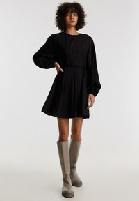 Schlichtes schwarzes Kleid mit Rundhalsausschnitt und Ballonärmeln, ausgestattet mit einem Taillengürtel und einem ausgestellten Rock; das Modell trägt graue knielange Stiefel.