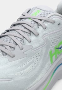 Chaussure de sport grise en tissu maille, avec un bout arrondi et une languette rembourrée. Comprend un logo vert et des accents verts, des lacets texturés et une semelle amortissante.