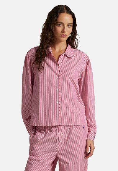 Femme aux longs cheveux bouclés portant un pyjama à rayures roses et blanches avec des boutons, debout avec une main dans la poche devant un fond blanc.