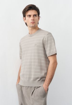 Νέος άνδρας που φοράει μπεζ κοντομάνικο πουλόβερ με λεπτές λευκές ρίγες και γκρι παντελόνι, κοιτάζοντας ελαφρώς προς τα πλευρά με τα χέρια στις τσέπες.