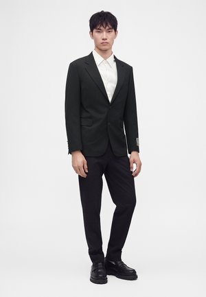 Jeune homme debout portant une veste de costume noire, une chemise blanche, un pantalon noir et des chaussures habillées noires sur un fond clair uni.