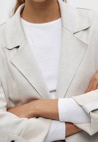 Personne aux bras croisés portant un blazer beige sur une chemise blanche côtelée, visible du cou à la taille.