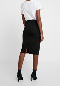 Jupe crayon noire en matière extensible, dotée d'une fente à l'arrière pour faciliter les mouvements. Portée avec un t-shirt blanc ajusté et des bottines noires à talons.