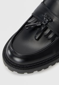 Mocasín de cuero negro con un acabado suave, que presenta un adorno de flecos y un lazo. Suela de goma gruesa con bordes texturizados.