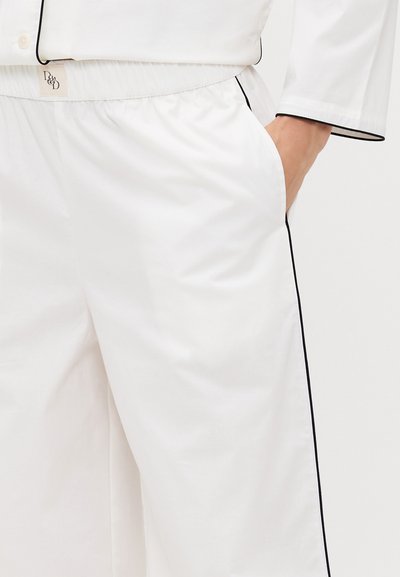 Pantaloni de pijama albi cu talie elastică, buzunare laterale și piping negru în contrast de-a lungul cusăturilor. Textura netedă a țesăturii este vizibilă.