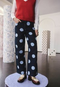 A sötét háttérrel rendelkező farmer széles lábú nadrág világos kék pöttyökkel van díszítve. Piros pulóverrel és két színű cipővel párosítva.