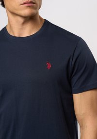 Tamno plava pamučna majica s okruglim izrezom, s malim crvenim vezom logotipa na lijevoj strani grudi. Glatka tekstura, kratki rukavi.