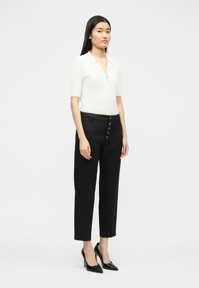 Γυναίκα όρθια, φοράει λευκή μπλούζα με κοντά μανίκια και γιακά, και μαύρο ψηλόμεσο παντελόνι με κουμπιά, συνδυασμένα με μαύρα τακούνια.