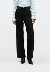 Μαύρα τζιν παντελόνια σε φαρδιά γραμμή, κατασκευασμένα από τζιν με ψηλή μέση. Διαθέτουν μπροστινές τσέπες και �κλείσιμο με κουμπί. Συνδυάζονται με μπλε μπλούζα με ρίγες.