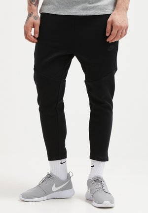 Uomo che indossa pantaloni della tuta neri a gamba stretta, calzini bianchi Nike e scarpe da ginnastica grigie Nike, in piedi su uno sfondo bianco uniforme.
