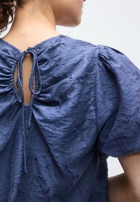 Μπλούζα σε μπλε χρώμα με υφή, που διαθέτει πλάτη με ραμπούλες και λεπτομέρεια κορδονιού. Απαλό, ζαρωμένο ύφασμα και φουσκωτά μανίκια δημιουργούν μια χαλαρή σιλουέτα.