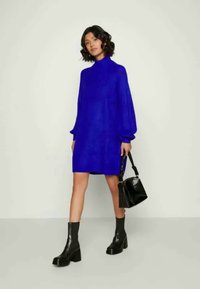 Royal kék pulóverruha magas nyakkal, hosszú puffos ujjakkal és textúrált anyaggal. Párban vastag fekete bokacsizmákkal és egy fekete kézi táskával.