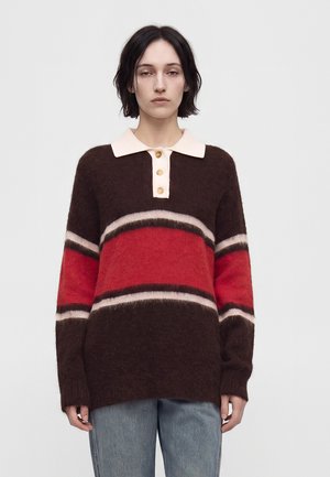 Eine junge Person mit kurzen dunklen Haaren trägt einen braun-roten und beigen gestreiften Pullover mit einem beigen Kragen und drei Knöpfen, kombiniert mit blauen Jeans.