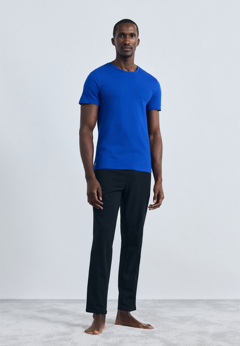 T-shirt di cotone blu con collo tondo, abbinata a pantaloni neri. Il modello si trova su un morbido tappeto grigio, a piedi nudi. Design semplice, senza motivi.