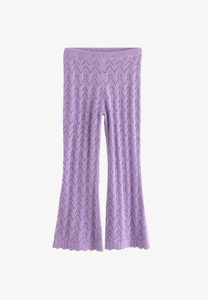 Ung pige med krøllet hår iført matchende lavendelfarvet strikket top og bukser, stående med hænderne bag ryggen mod en ensfarvet baggrund.