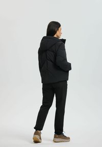 Frau in schwarzer Kapuzensteppjacke, schwarzen Hosen und beigen Wanderschuhen, die steht und nach rechts schaut, vor einem weißen Hintergrund.